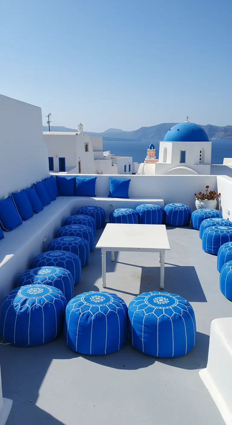 Bright blue poufs and cushions on a whitewashed rooftop overlooking the sea and a blue-domed church.