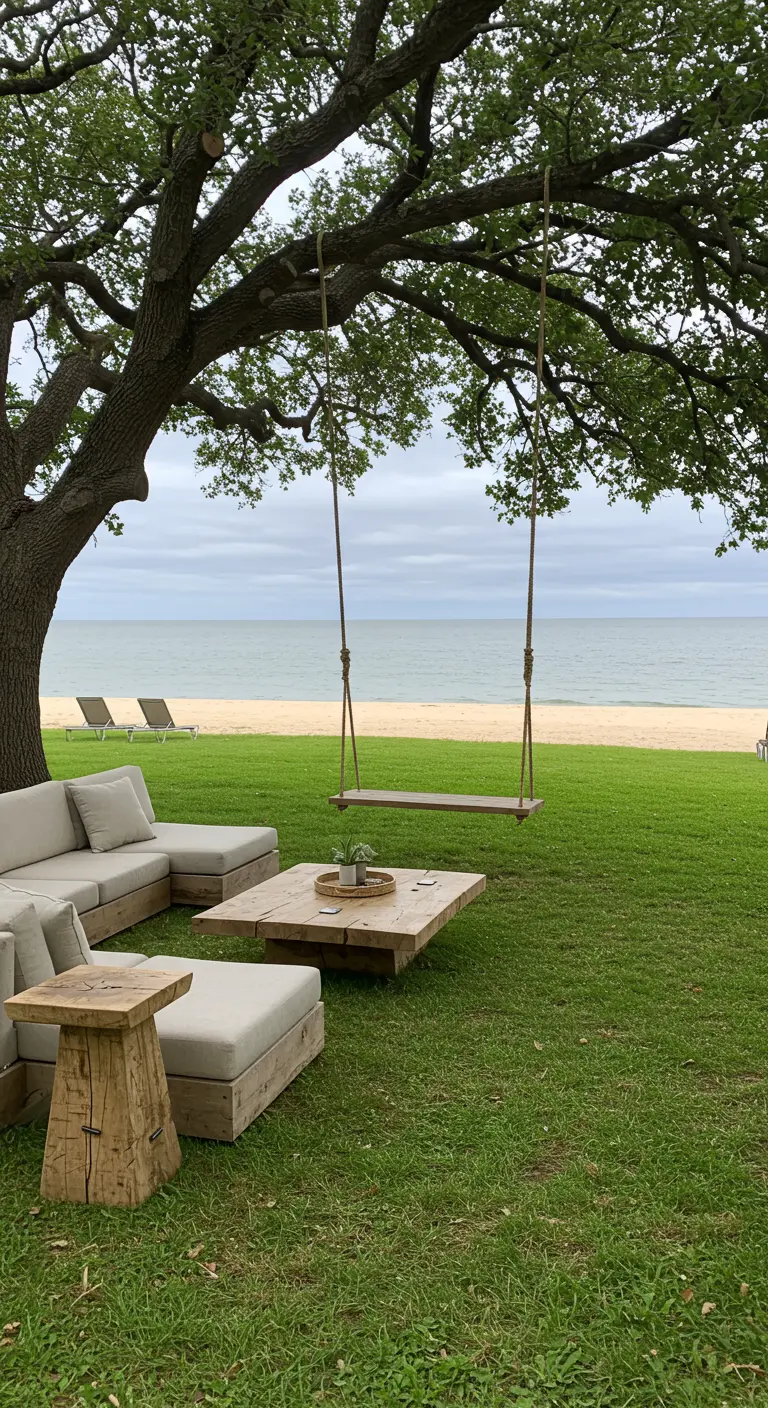 A large tree on a lawn by the sea, with a rope swing, sectional sofa, and driftwood table.