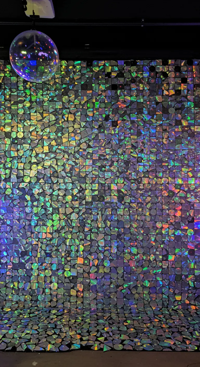 A backdrop wall covered in a mosaic of broken, iridescent CDs, creating a rainbow shimmer.