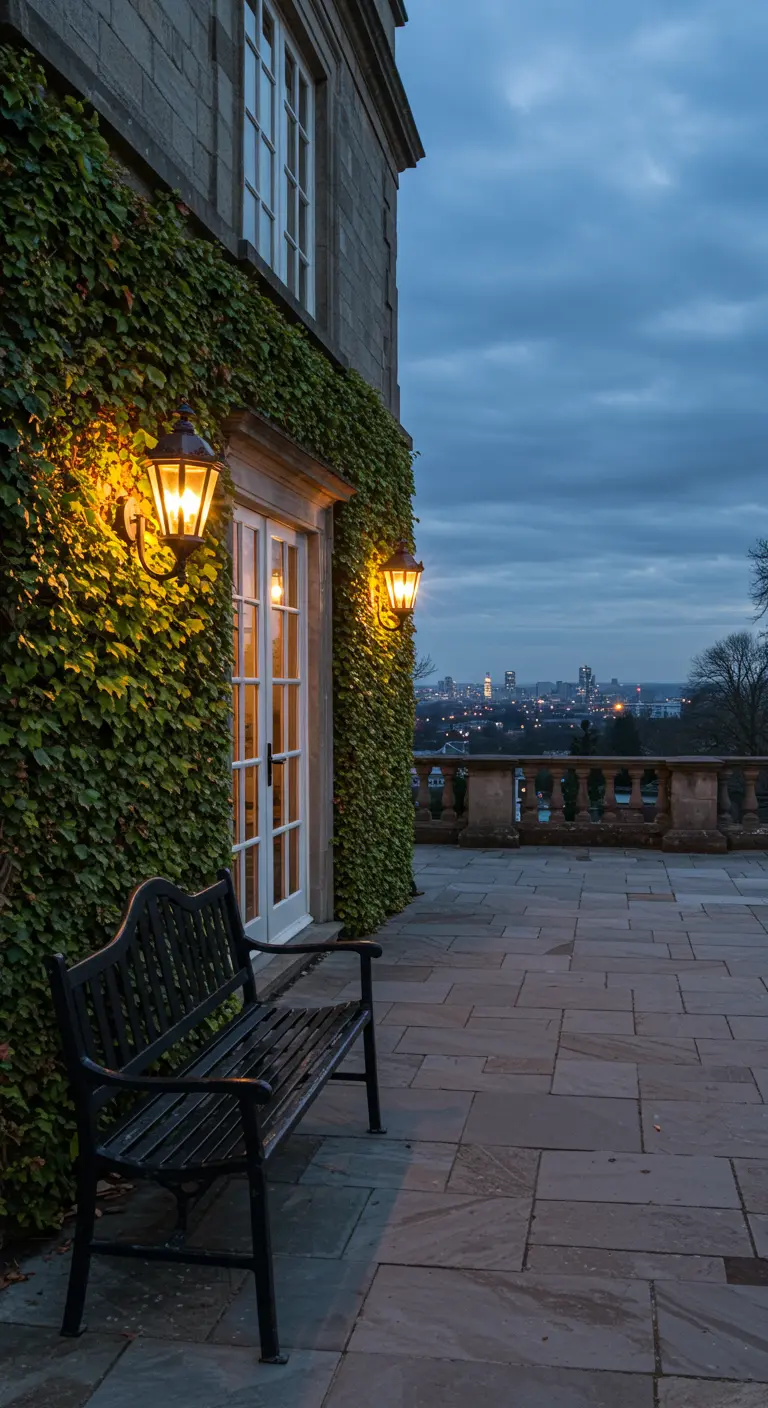 A black park bench on a stone terrace with an ivy-covered wall, overlooking a city skyline at dusk.