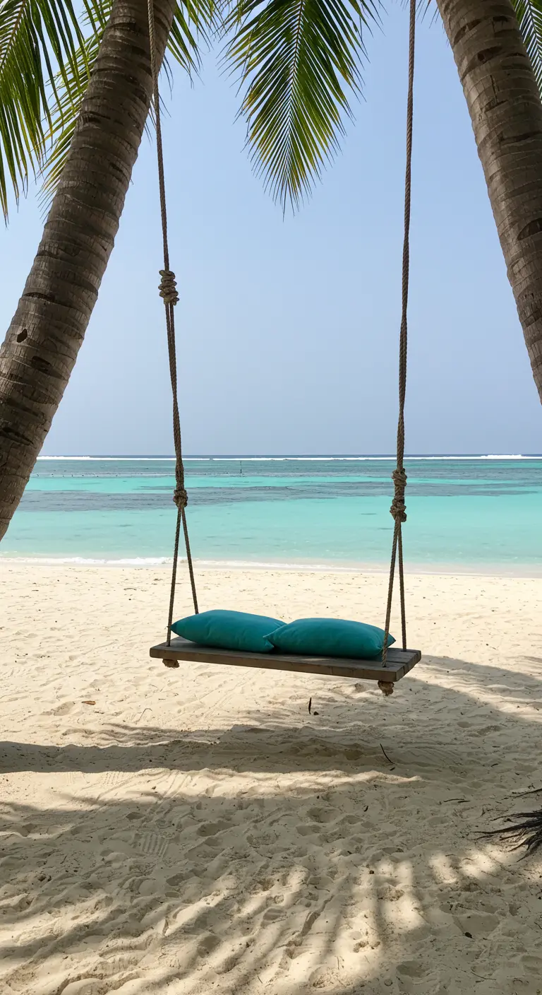 A simple wooden plank swing with two blue pillows hanging between two palm trees on a white sand beach.